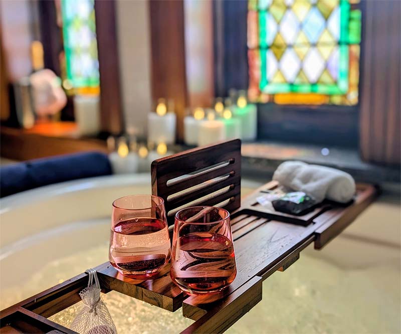 Candlelight spa experience — two glasses of wine resting on a bath tray with candles and stained glass in the background