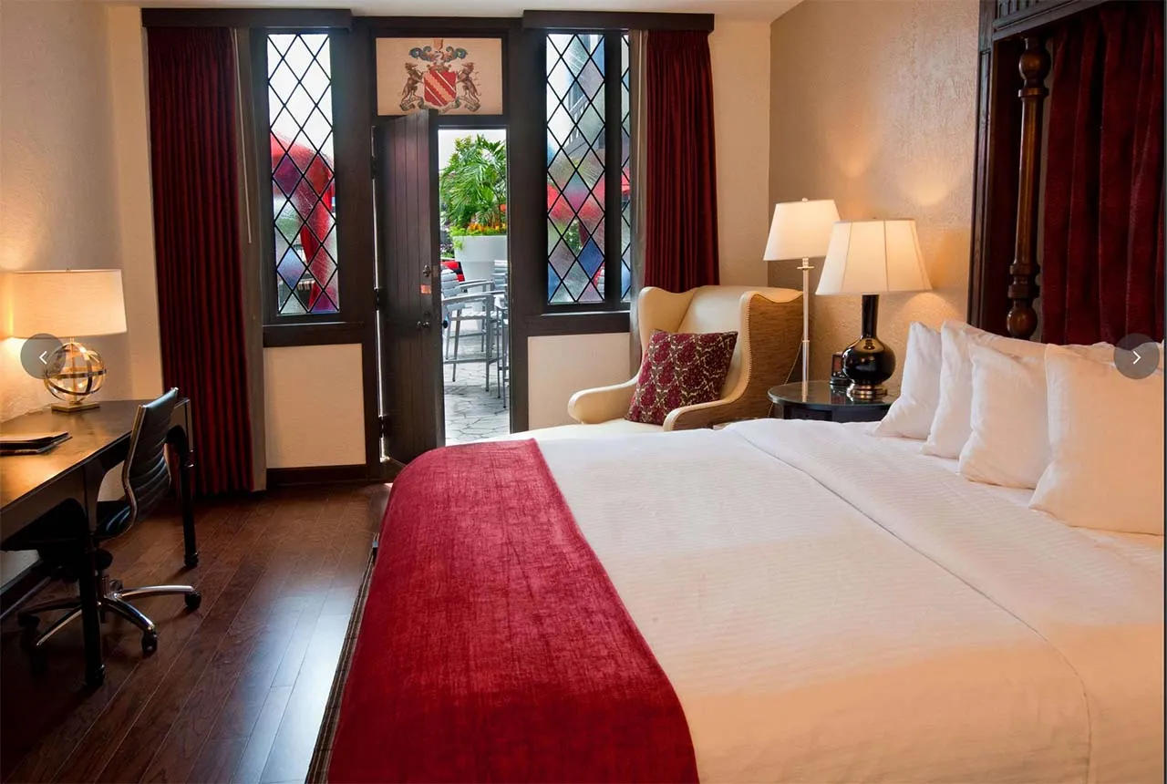 A guest room at The Cheshire Inn with a four-poster bed, crimson accents, leaded stained glass doors opening to a private patio, and dark wood furnishings