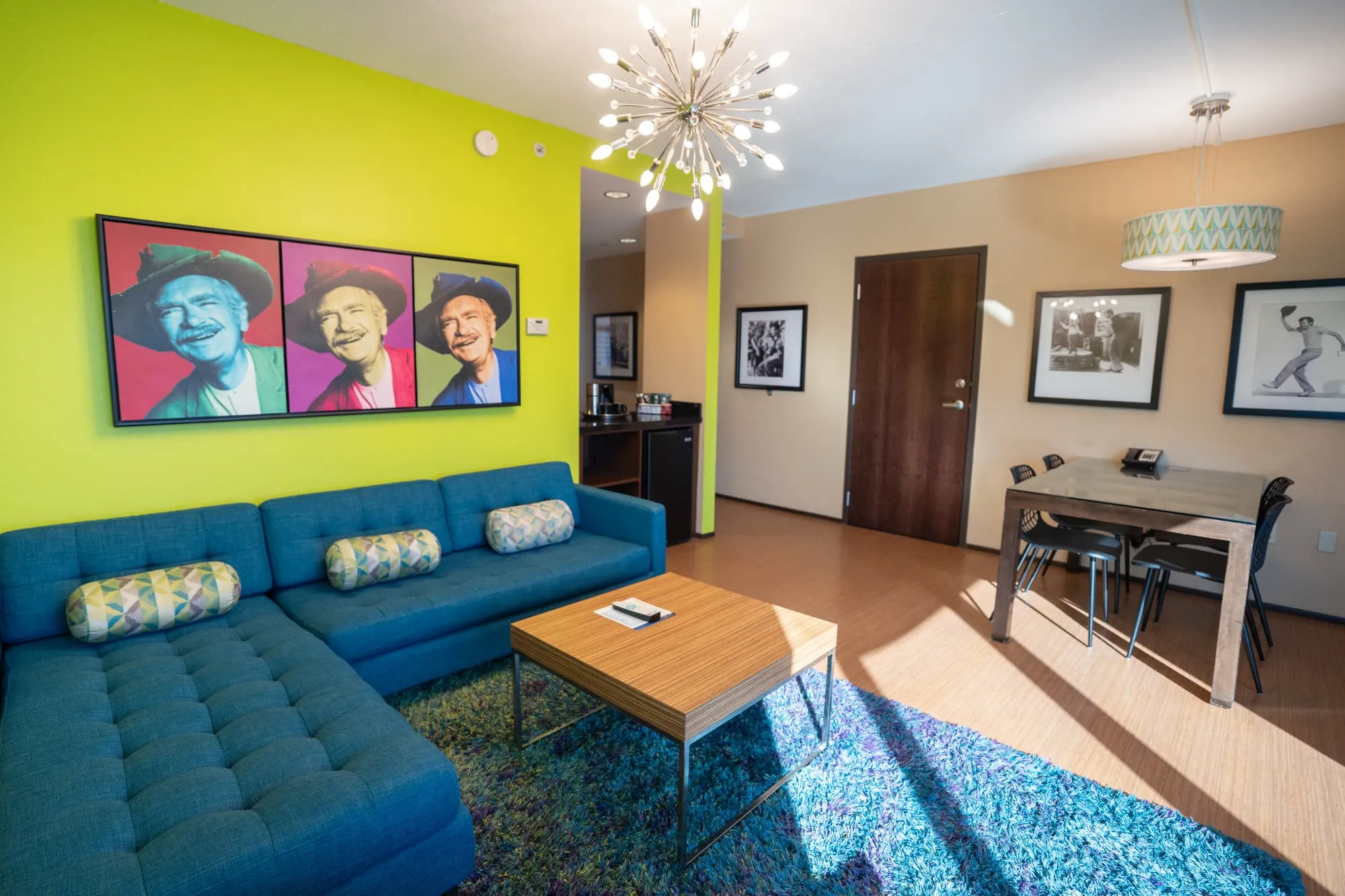 A Walk of Fame celebrity suite at The Moonrise Hotel featuring a chartreuse accent wall, Warhol-style portrait, teal sectional sofa, and dining table for four